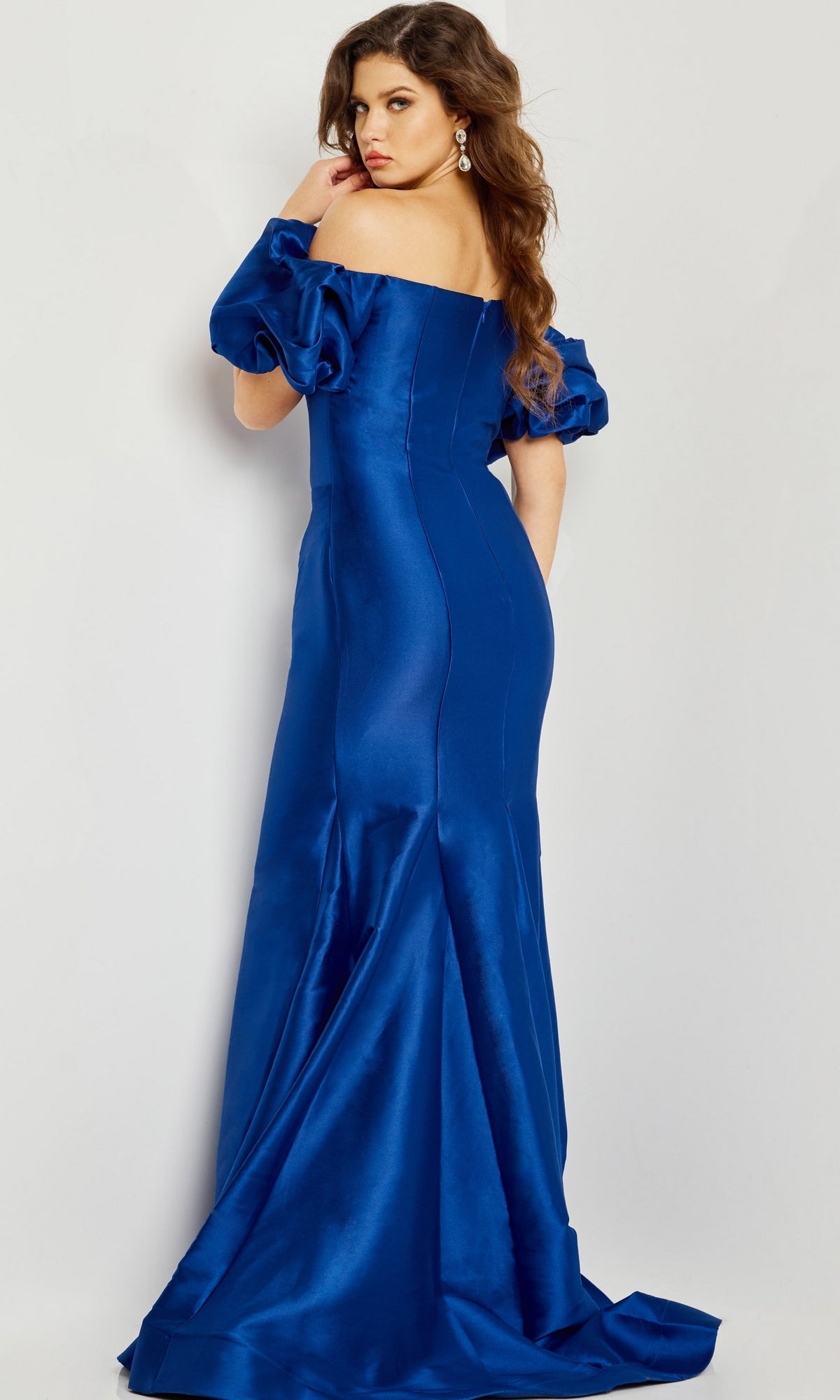 A woman wears the Jovani 08361 Puff-Sleeve Mikado Evening Dress in shiny royal blue, featuring off-the-shoulder puffed sleeves and a mermaid silhouette, posing slightly turned away against a plain background.